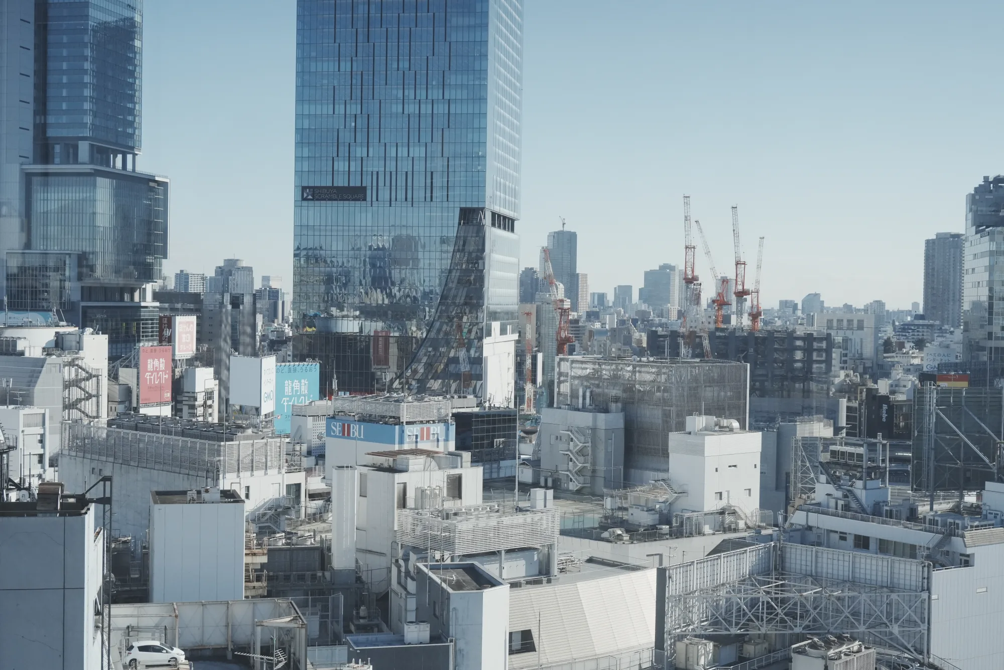 Glass towers and construction cranes rise above a dense Tokyo skyline under a pale blue sky.